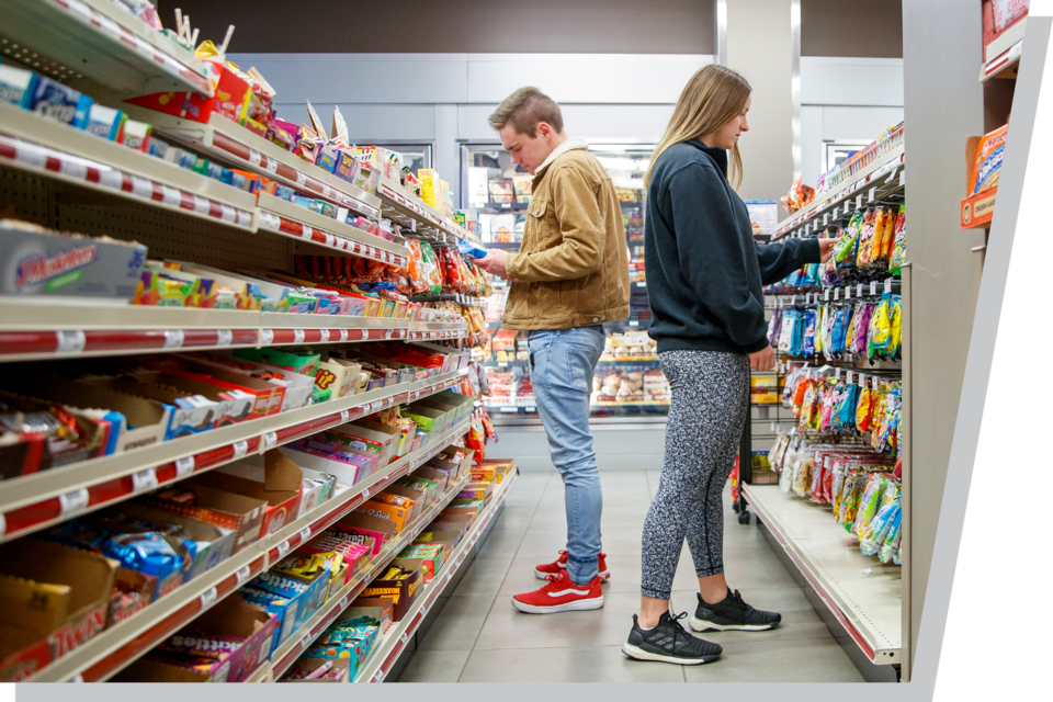 Two people browsing snack aisles in a well-lit convenience store.
