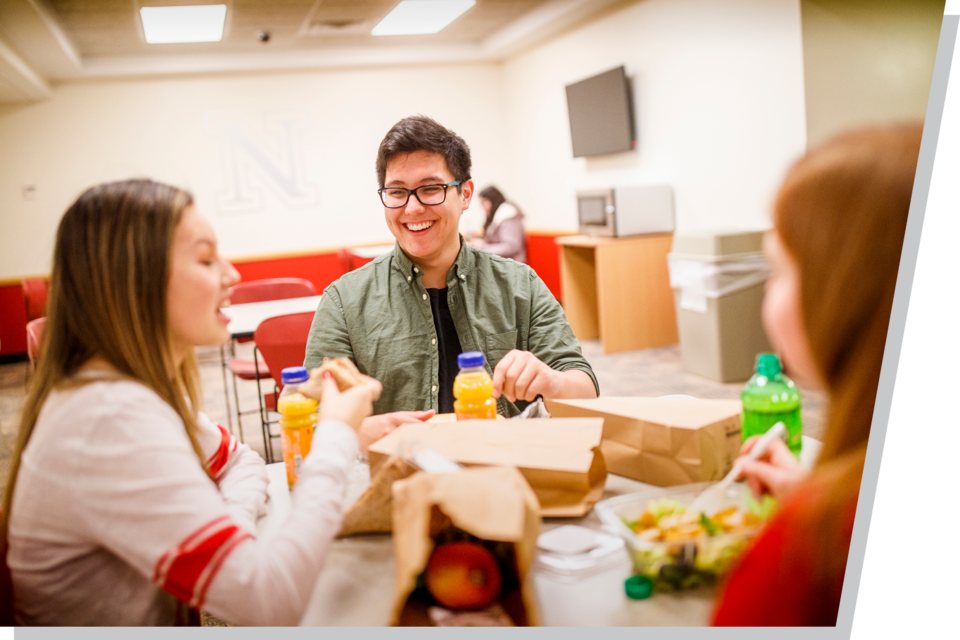 A male student and two female students sit around a table, with their sack lunches out, eating lunch in a cafeteria.