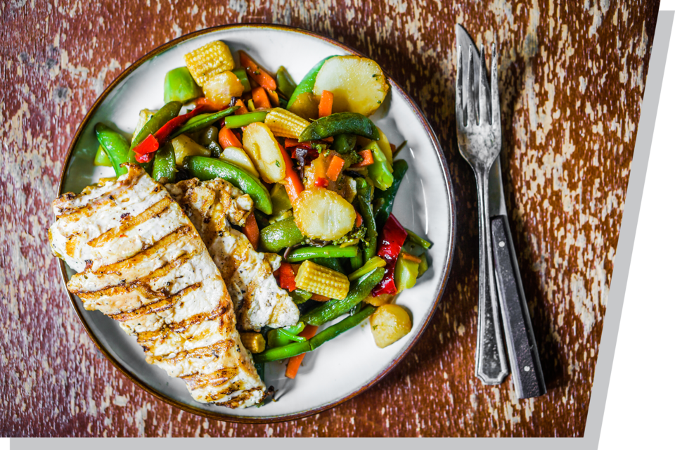 Grilled chicken with mixed vegetables on a plate, resting on a rustic wooden table alongside silverware.