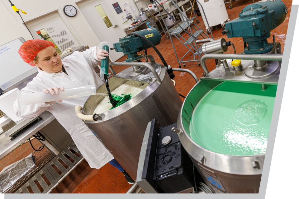 Worker in lab coat adds green liquid to industrial mixing tank in factory setting.