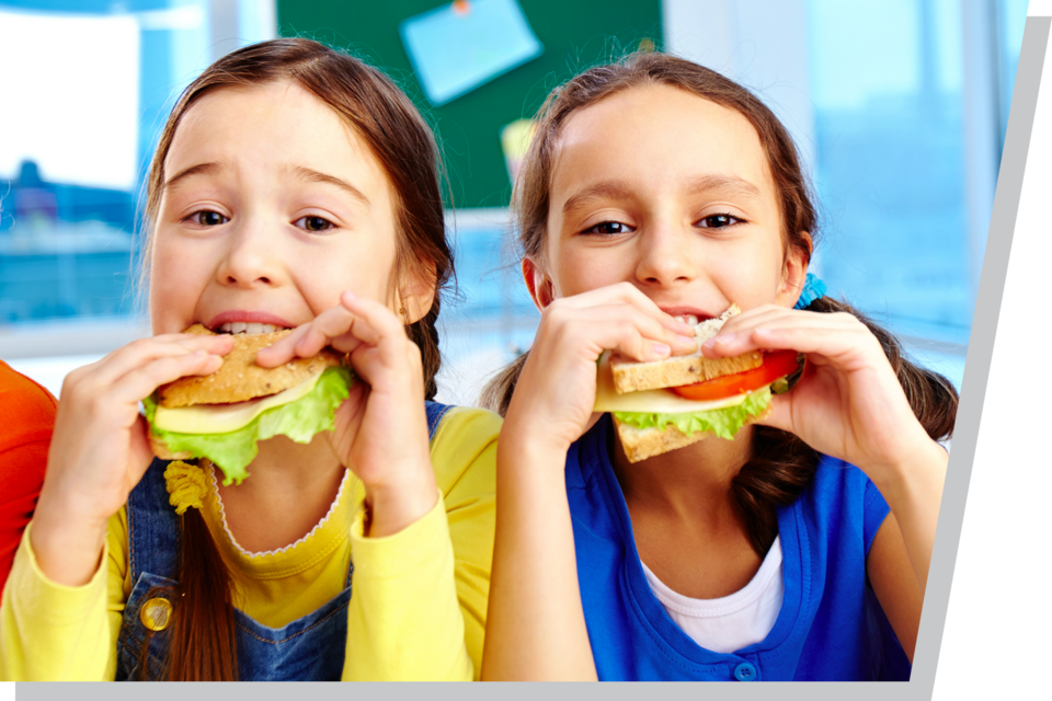 Two children eating sandwiches with lettuce and tomato in a bright room.